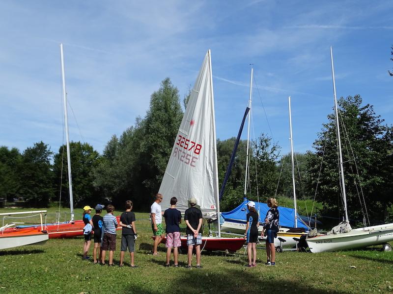 Einige Jugendliche stehen vor den Segelbooten und einem Lehrer im Segelcamp, welcher währenddessen erklärt