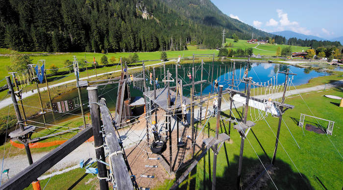 Teilnehmer eines Sommercamps für Kinder klettern durch einen Hochseilgarten an einem See, der von Bergen umgeben ist