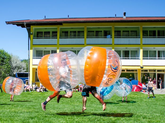 Eine Gruppe Jugendliche spielt Bubble Soccer auf einer grossen Wiese vor dem Ferienhaus im Segelcamp.