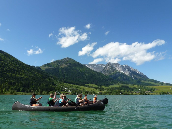 Jugendliche sitzen in Kanus auf einem See umgeben von einem Bergpanorama im Sportlager.