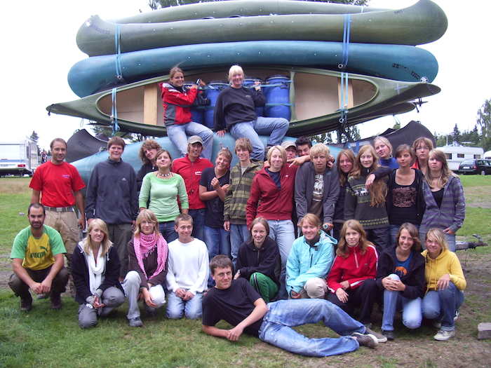 Eine Gruppe Kinder und Betreuer vom Kanu-Camp stehen vor einer Menge aufgestapelter Kanus.