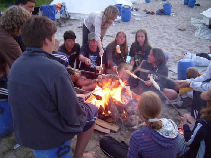 Eine Gruppe Kinder Kanu-Camp sitzt mit Stöcken am Lagerfeuer im Sand.