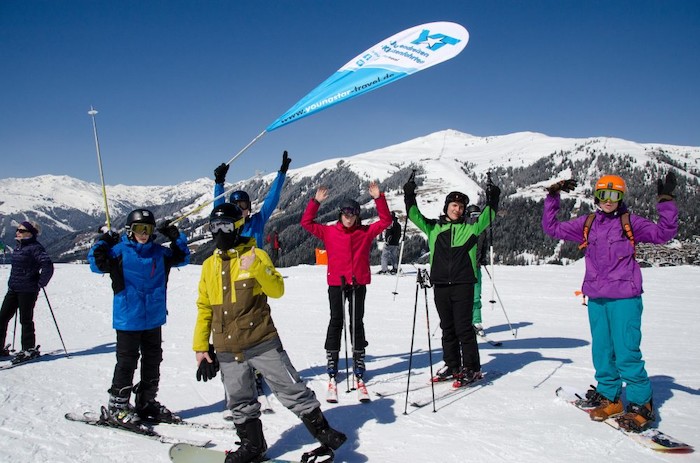 Eine Gruppe Jugendlicher steht am Pistenanfang und hält die Arme mitsamt Ski Stöcken in die Höhe im Skilager.
