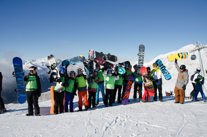 Eine Gruppe Jugendlicher steht am Pistenanfang und hält ihre Snowboards in die Höhe im Skilager.