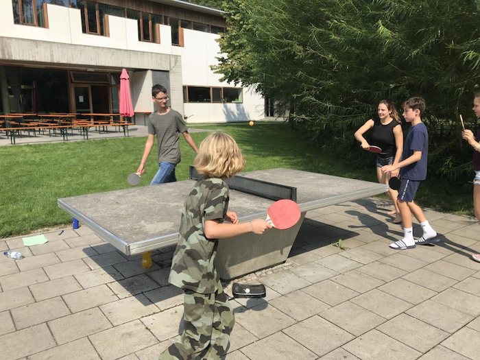 Die Teilnehmer eines Lerncamps Sommerferien spielen gegeneinander Tischtennis und laufen um im Kreis um die Platte herum