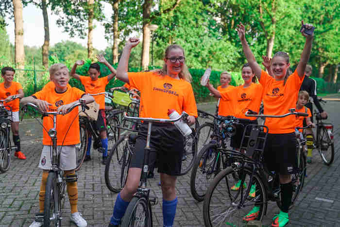 Teilnehmer eines Sport-Camps stehen mit Fahrrädern auf einer Straße und halten an