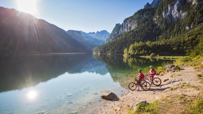 Zwei Jungs stehen mit ihren Mountainbikes am Seeufer, in dem sich die Berge spiegeln während ihrer Sommerfreizeit Kinder
